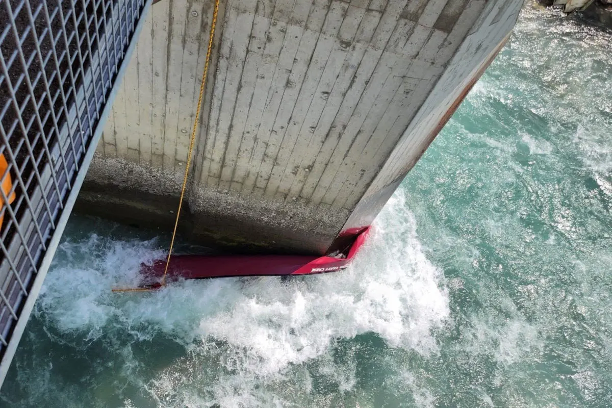 Rote Schutzbarrieren sind an einer Betonbrücke befestigt. Unterhalb fließt turbulent blaues Wasser.