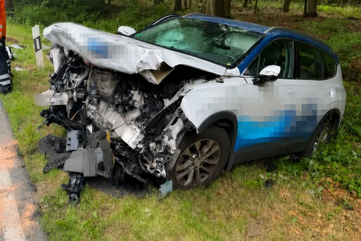 Ein stark beschädigtes weißes Auto steht auf einer Wiese neben einer Straße. Die Front ist komplett eingedrückt.