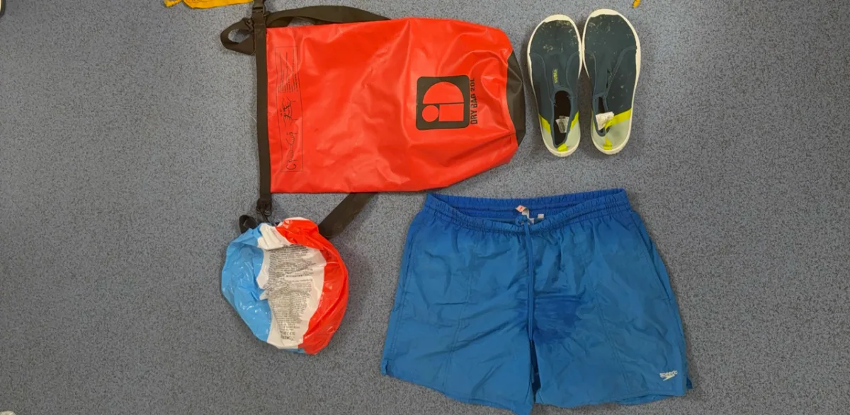 Ein roter wasserdichter Seesack mit schwarzem Logo, ein Paar blaue Aquaschuhe, eine blau-weiße Badehose und ein in Rot-Weiß-Blau verpackter Gegenstand liegen auf grauem Teppichboden.