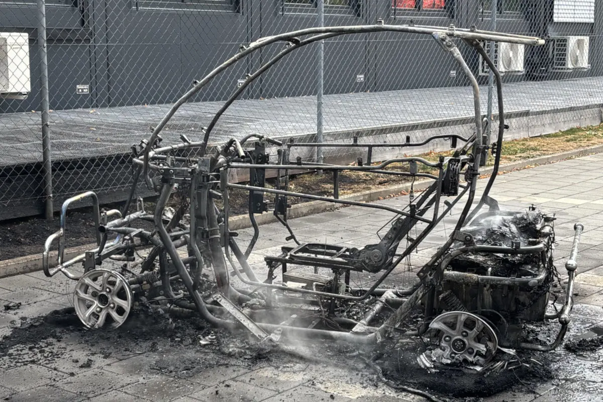 Ein vollständig ausgebranntes Fahrzeuggestell steht auf einem Gehweg. Rauch steigt noch aus dem Wrack auf.