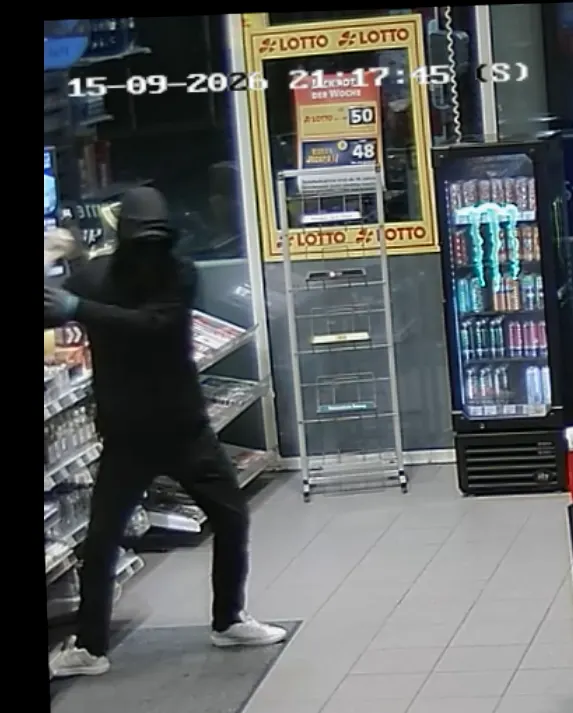 Ein maskierter Mann mit schwarzer Kleidung und einer Kapuze hält eine Waffe in einem Geschäft. Im Hintergrund sind Lottowerbung und Getränkeautomaten zu sehen.