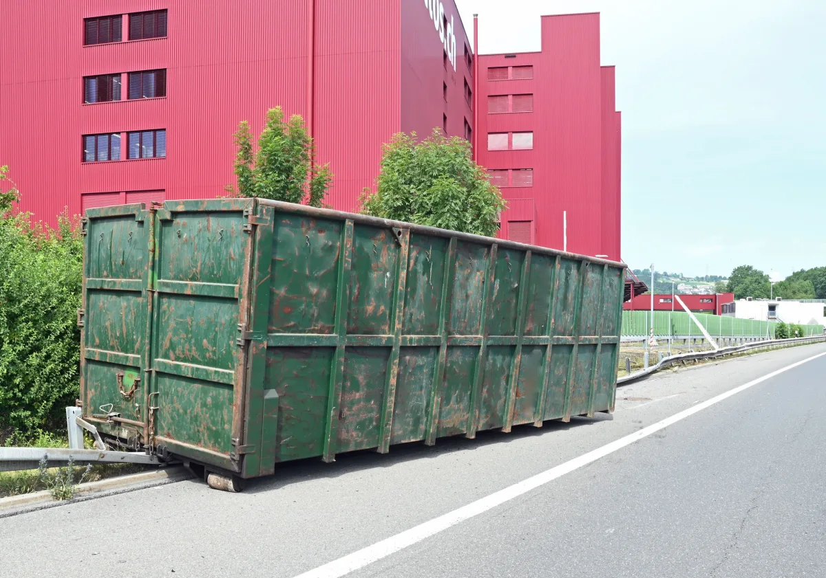 Ein grüner Container liegt auf der Seite einer Straße, nahe einem großen, roten Gebäude. Im Hintergrund sind auch grüne Bäume sichtbar.