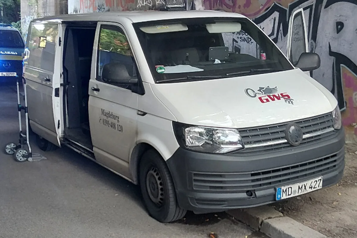 Ein weißer Transporter steht mit geöffneten Vordertüren neben einem Polizeiauto unter einer bemalten Brücke.