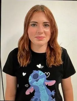Eine junge Frau mit schulterlangem Haar trägt ein schwarzes T-Shirt mit einem Comic-Aufdruck und steht vor einer neutralen Wand.