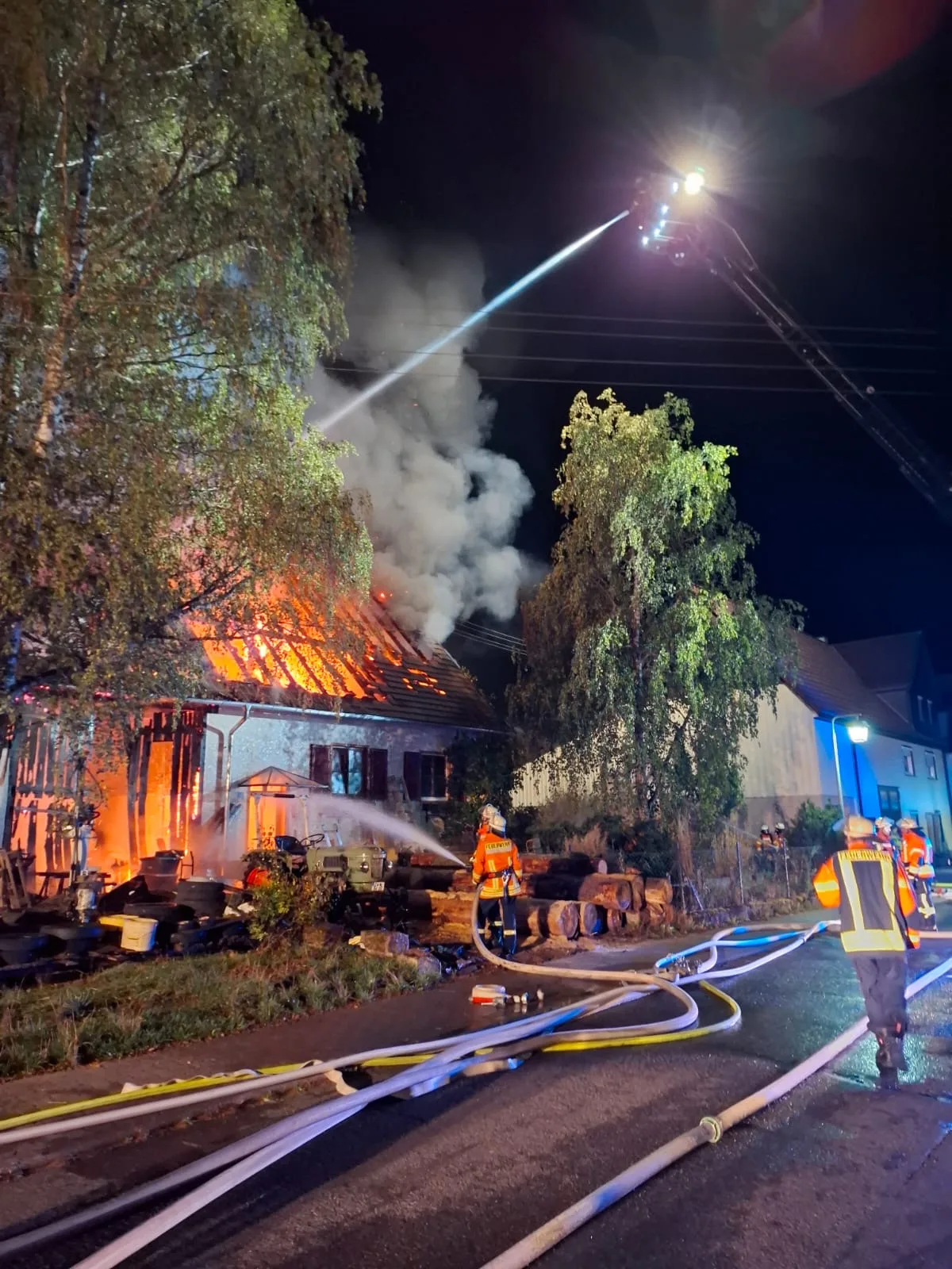 Mehrere Feuerwehrleute bekämpfen bei Nacht den Brand eines Wohnhauses. Flammen schlagen aus dem Dach, während Wasserschläuche zum Einsatz kommen.