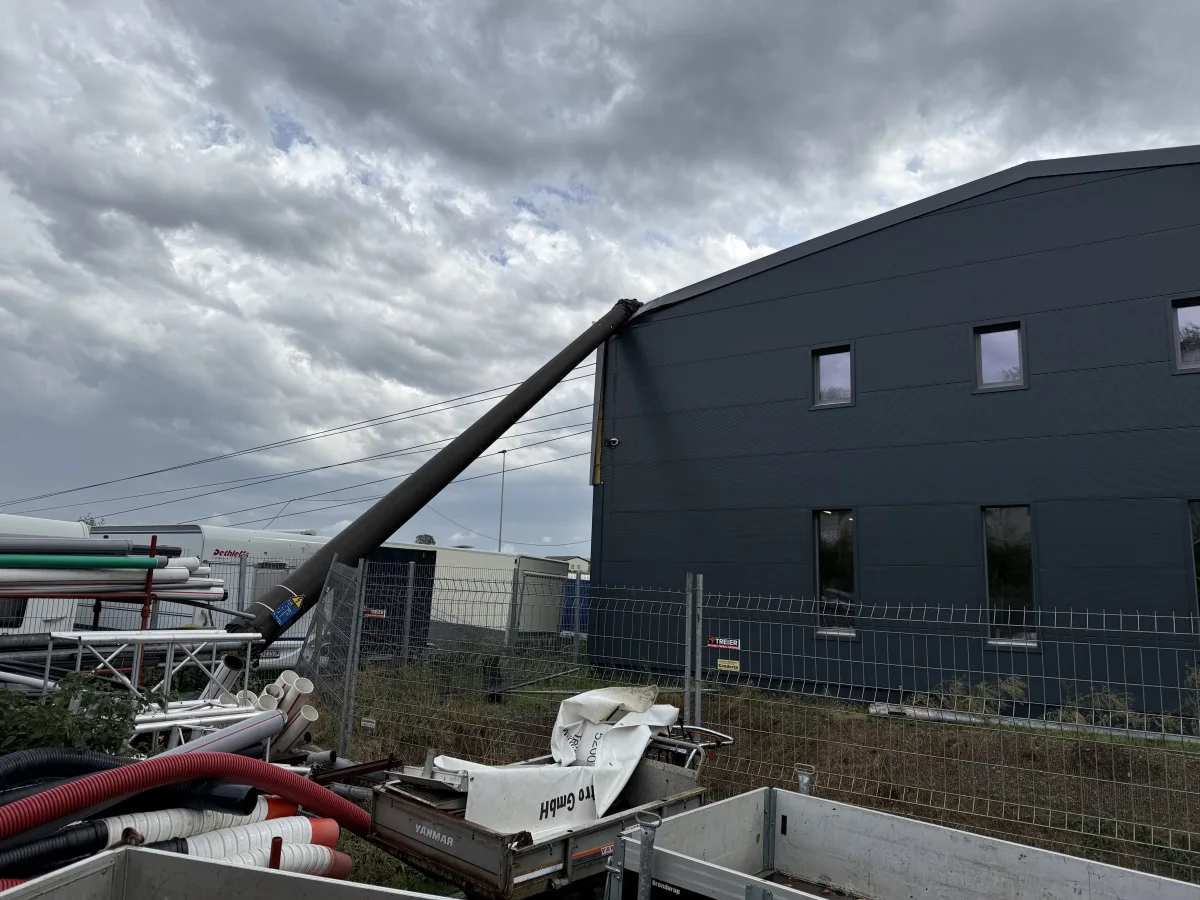 Ein Strommast neigt sich und lehnt gegen ein dunkles Gebäude. Am Boden liegen Rohre und Baustellenmaterialien unter einem wolkigen Himmel.