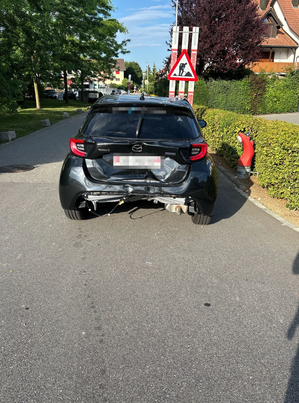 Ein schwarzer Mazda parkt am Straßenrand, das Heck zeigt deutliche Beschädigungen. Im Hintergrund sind Häuser und Straßenschilder zu sehen.