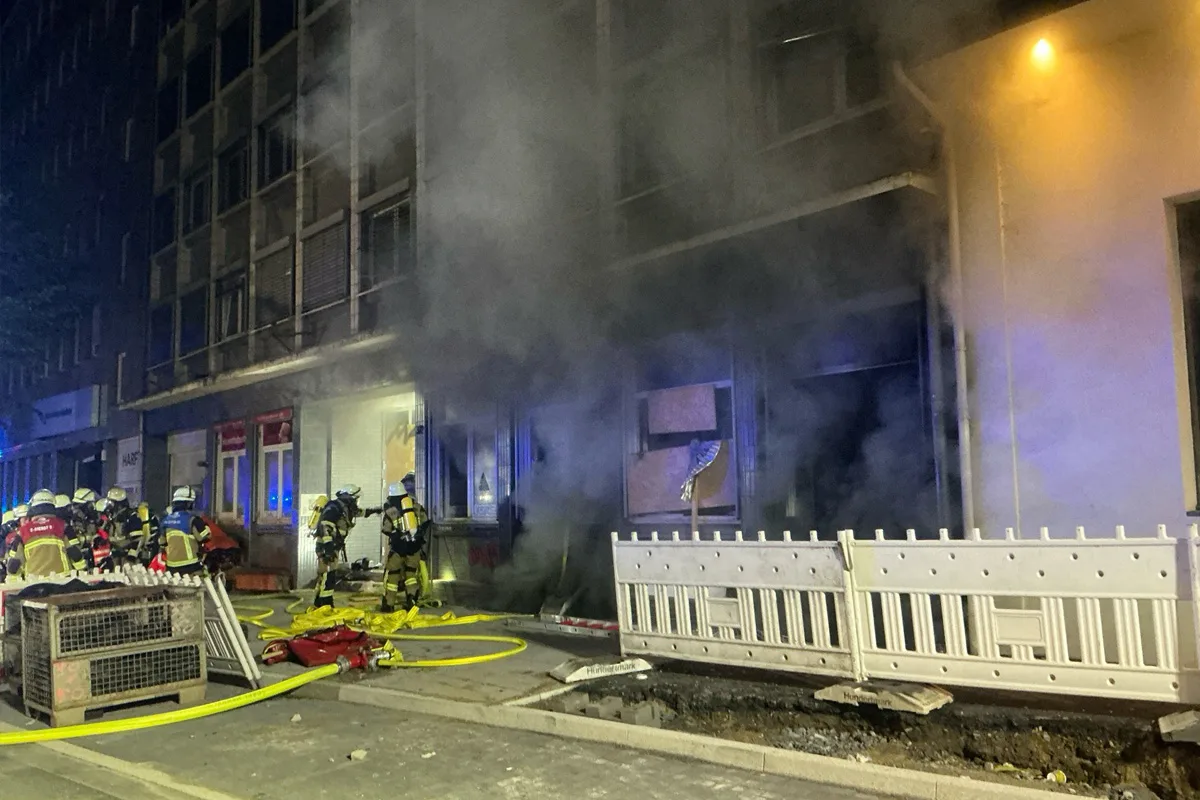 Mehrere Feuerwehrleute in Schutzausrüstung sind vor einem rauchenden Gebäude im Einsatz. Die Szene findet bei Nacht in einer städtischen Umgebung statt.