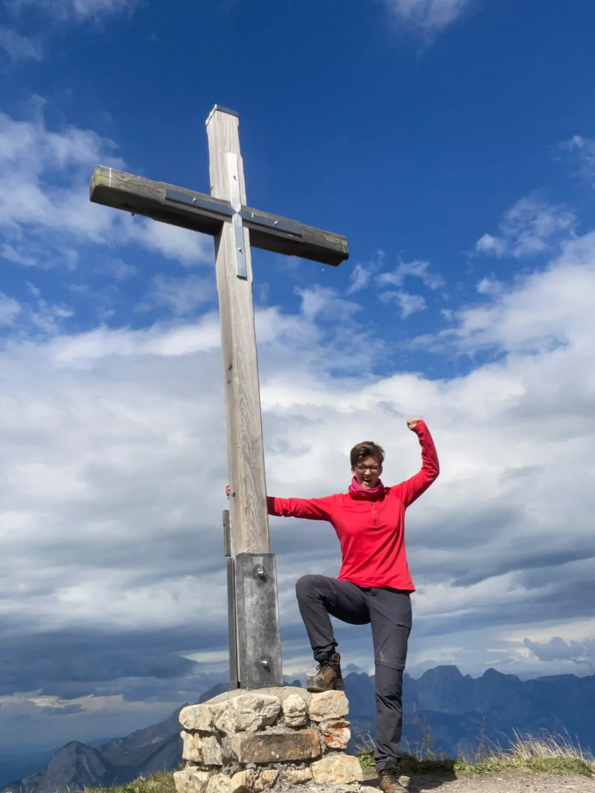 Eine Person in einer roten Jacke posiert neben einem Holzkreuz auf einem Berggipfel, während im Hintergrund Wolken am Himmel zu sehen sind.