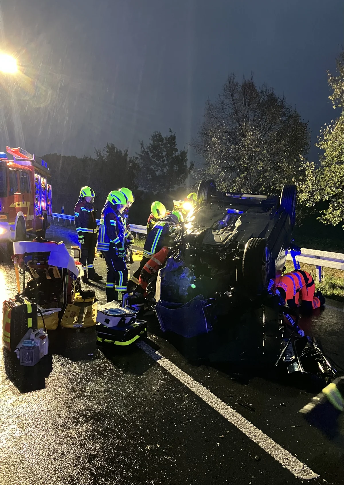 Mehrere Feuerwehrleute in reflektierenden Anzügen arbeiten an einem auf dem Dach liegenden Auto auf einer regennassen Straße. Ein Feuerwehrauto steht in der Nähe.