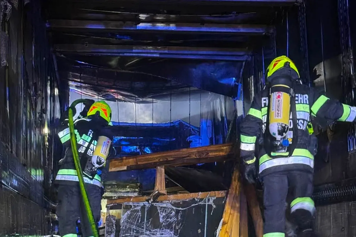 Zwei Feuerwehrleute in Schutzkleidung arbeiten in einem dunklen, verbrannten Innenraum. Einer hält einen Schlauch.
