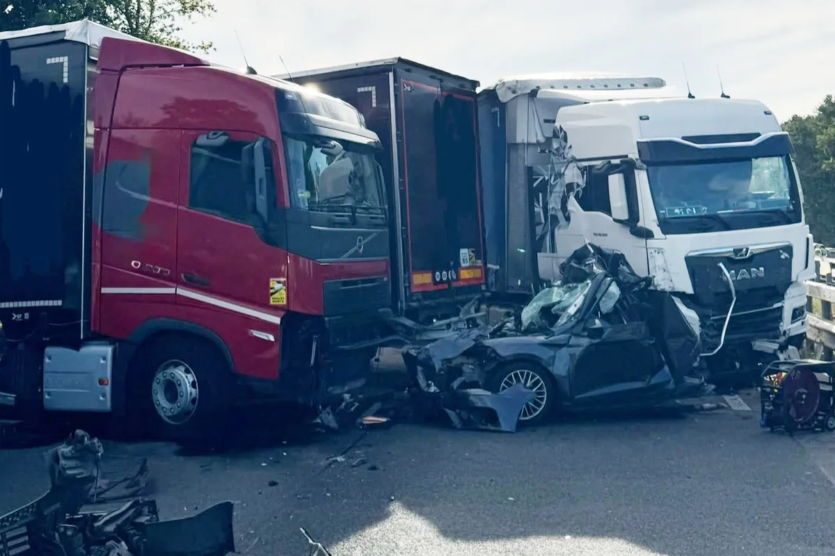 Ein schwarzer Pkw ist stark beschädigt und zwischen zwei Lkw eingeklemmt. Die Lkw sind rot und weiß, beide sichtbar beschädigt.