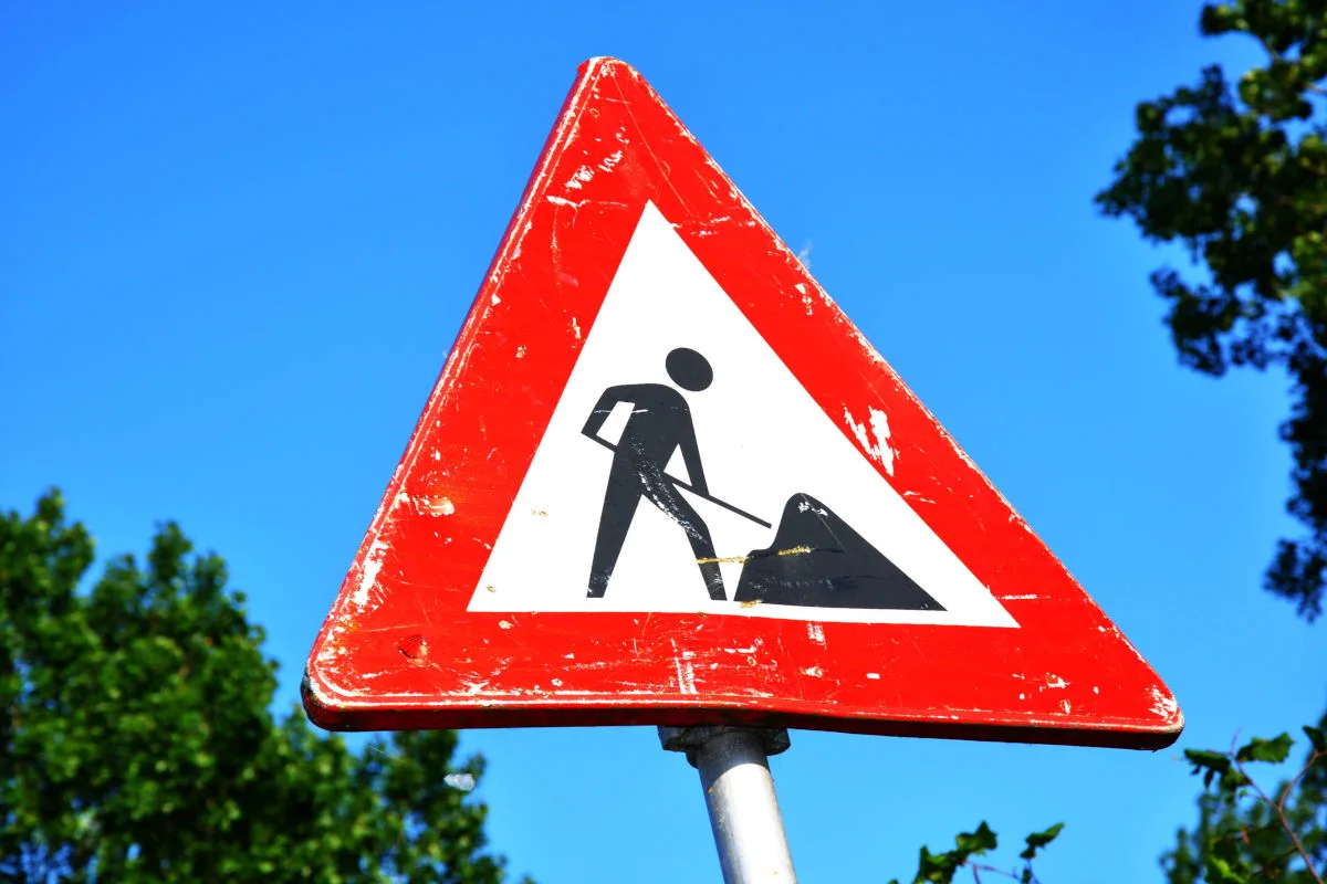 Ein dreieckiges Baustellenschild auf einer Straße, umgeben von Bäumen unter einem klaren blauen Himmel. Das Schild hat einen roten Rand und zeigt eine stilisierte Figur, die mit einem Spaten arbeitet.