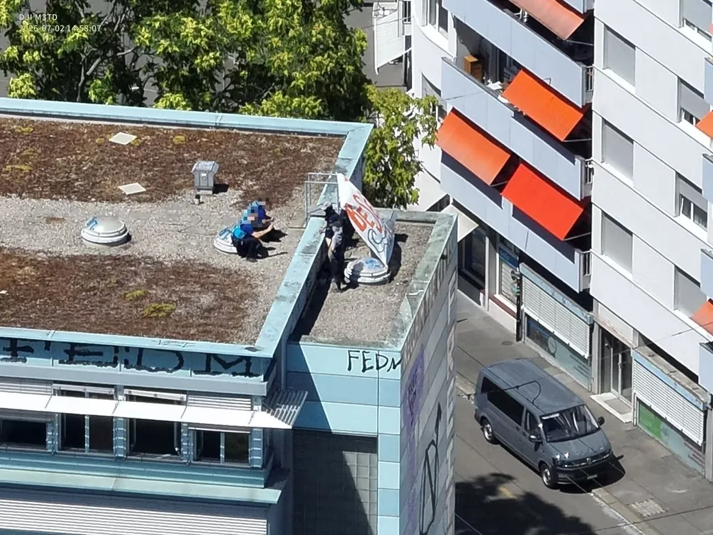 Zwei Personen sitzen auf dem Dach eines Gebäudes, das mit Kies bedeckt ist. Eine hat ein Schild in der Hand.