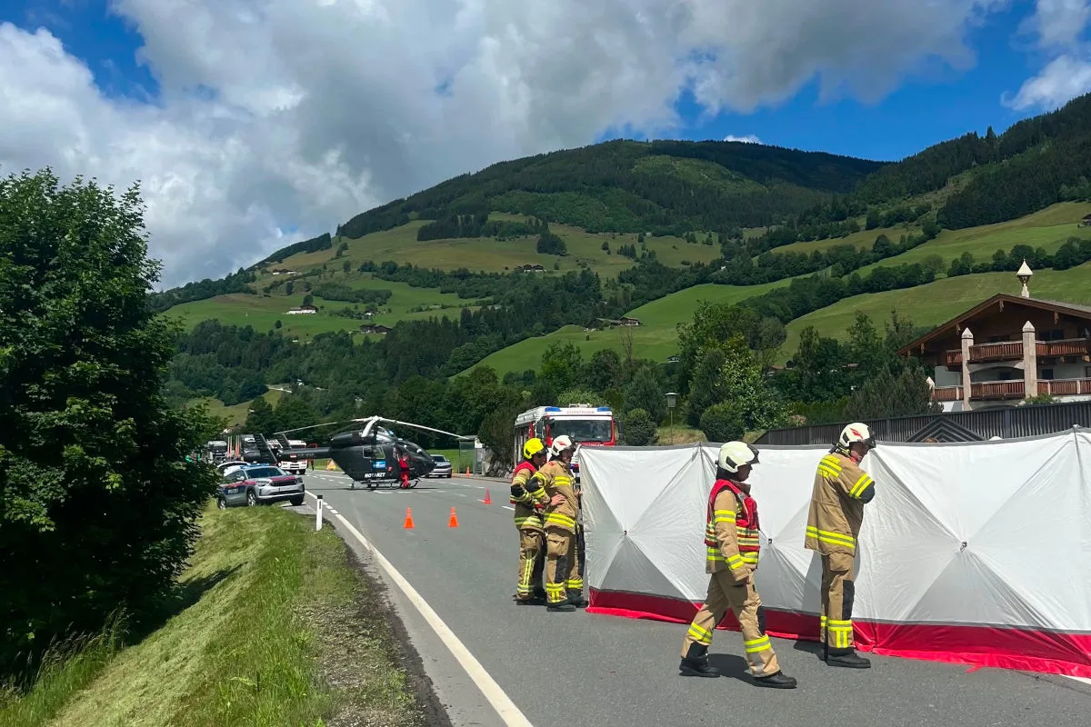 Ein Rettungshubschrauber steht auf einer Straße in einer grünen, bergigen Landschaft. Feuerwehrleute errichten eine Sichtschutzwand neben dem Unfallort.