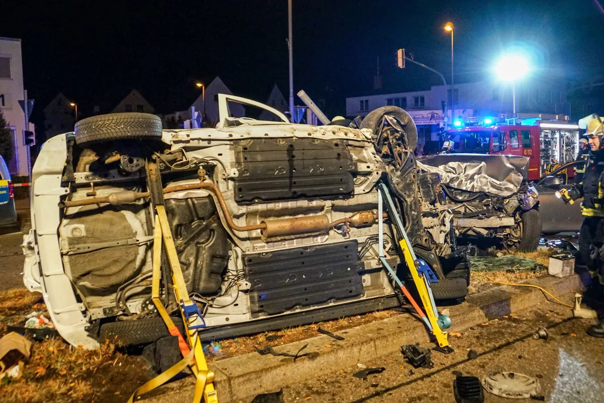 Ein Auto liegt auf der Seite, daneben ein weiteres stark beschädigtes Auto. Feuerwehrkräfte sind vor Ort und sichern die Stelle.