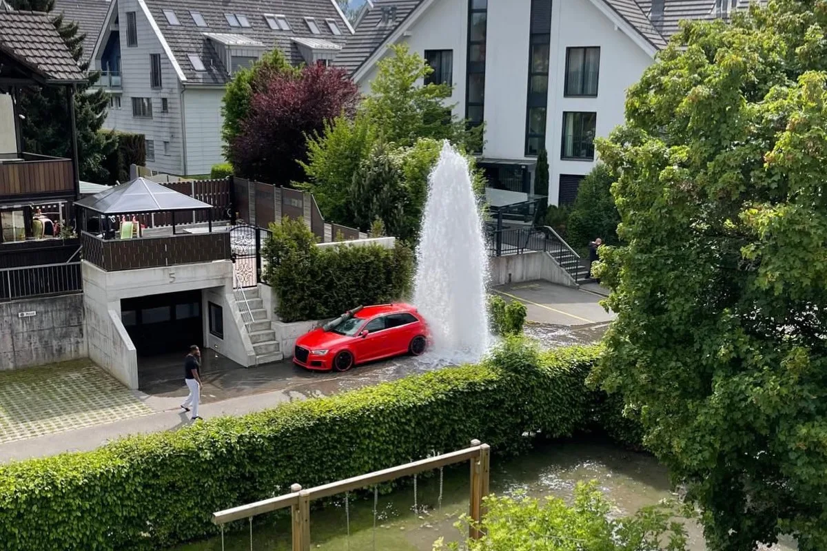 Ein rotes Auto steht in einem Wohngebiet neben einer großen Wasserfontäne, die aus dem Boden sprüht. Im Hintergrund sind Wohnhäuser und Bäume zu sehen.