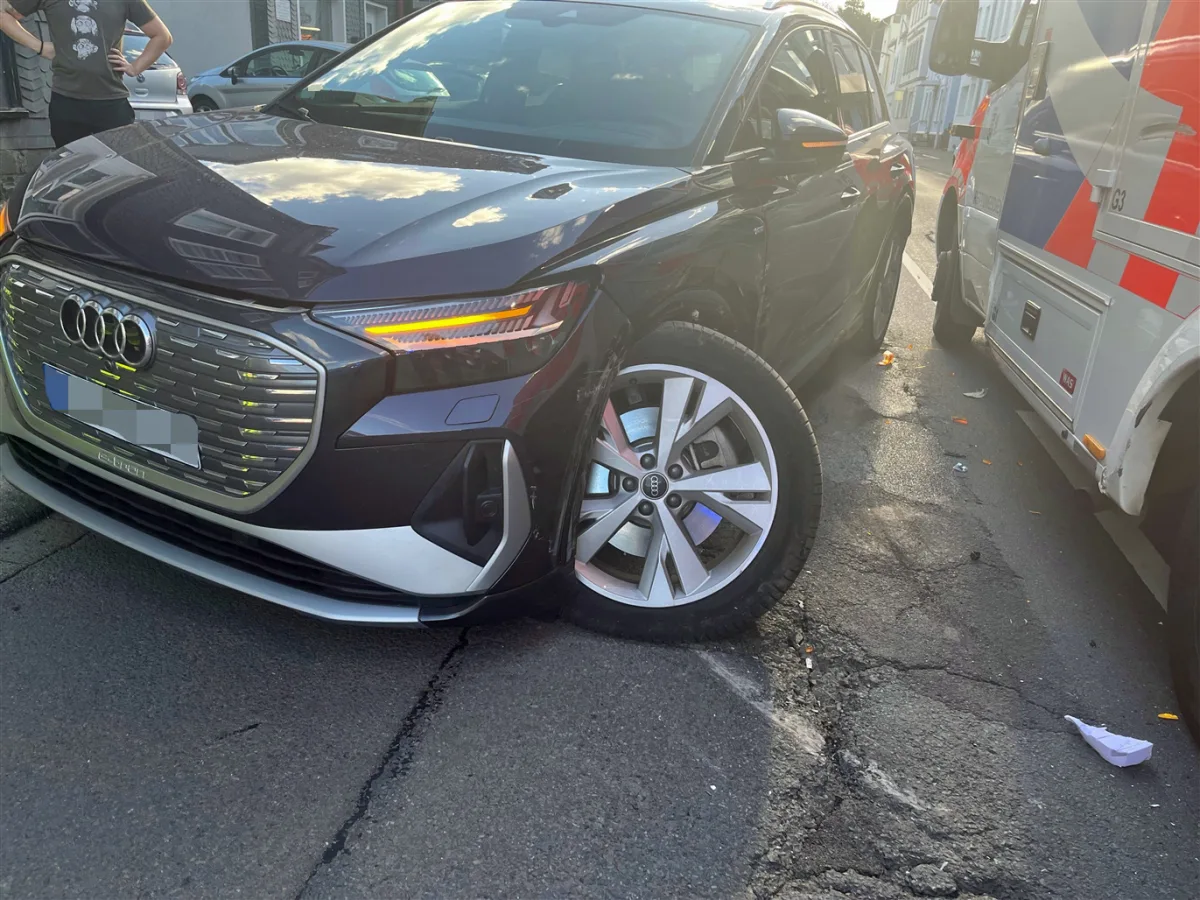 Ein schwarzer Audi mit sichtbarem Frontschaden ist auf einer Straße verkeilt mit einem Rettungsfahrzeug. Im Hintergrund sind Gebäude und eine Person zu sehen.