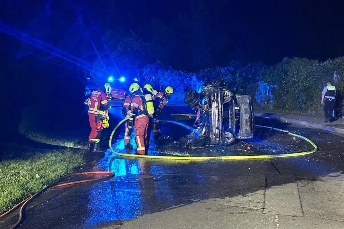 Mehrere Feuerwehrleute sind dabei, ein auf der Seite liegendes und brennendes Auto bei Nacht zu löschen. Blaulicht und Ausrüstung sind sichtbar.
