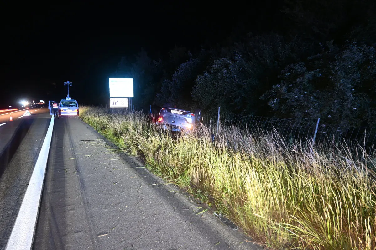 Ein Pkw liegt in einem Graben neben einer beleuchteten Autobahn. Ein Polizeifahrzeug steht auf dem Seitenstreifen mit Einsatzkräften.