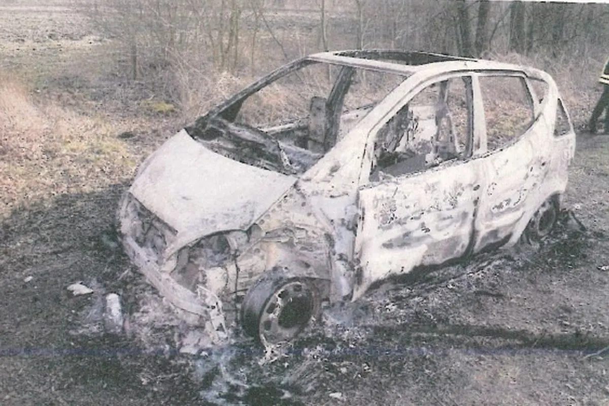 Ein stark ausgebranntes Auto steht auf einem unbefestigten Weg, umgeben von kahlen Bäumen. Das Fahrzeug ist vollständig zerstört.
