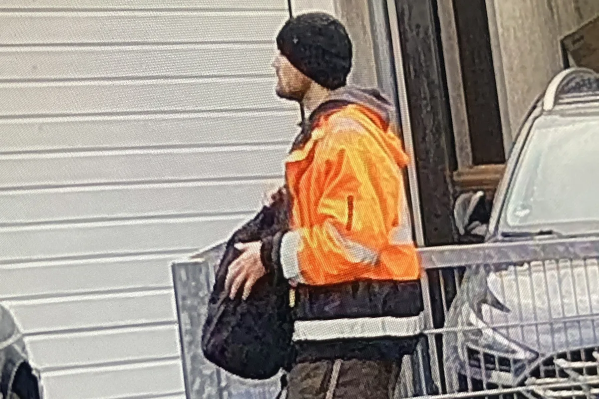 Ein Mann in einer leuchtend orangen Jacke und einer schwarzen Mütze hält eine Tasche. Er steht neben einem Auto vor einem Gebäude mit einer weißen Garagentür.