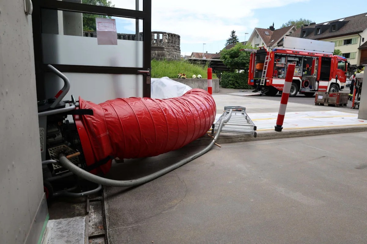Ein roter Lüftungsschlauch ragt aus einer geöffneten Tür in einen Innenhof; ein Feuerwehrfahrzeug steht bereit.