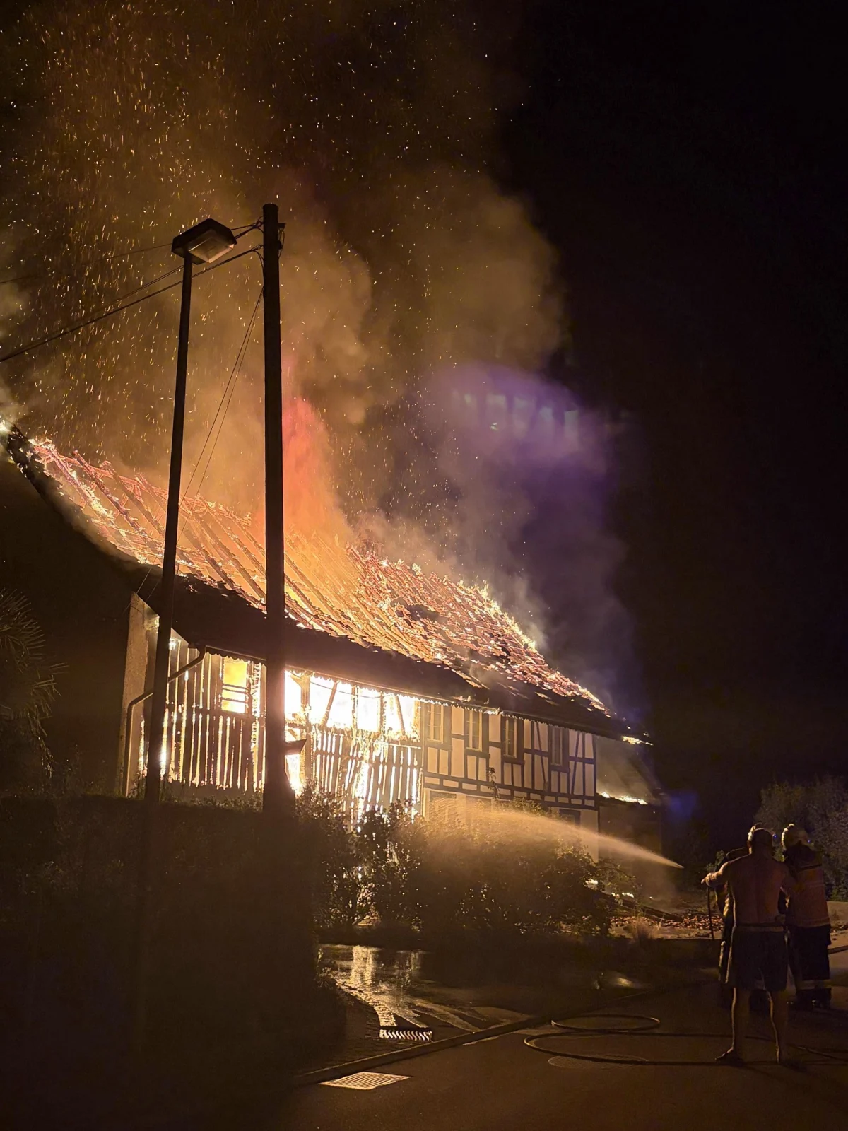Ein Fachwerkhaus ist in der Nacht in Brand geraten. Flammen schlagen aus dem Dach, und Feuerwehrleute sind im Einsatz, um das Feuer zu löschen.