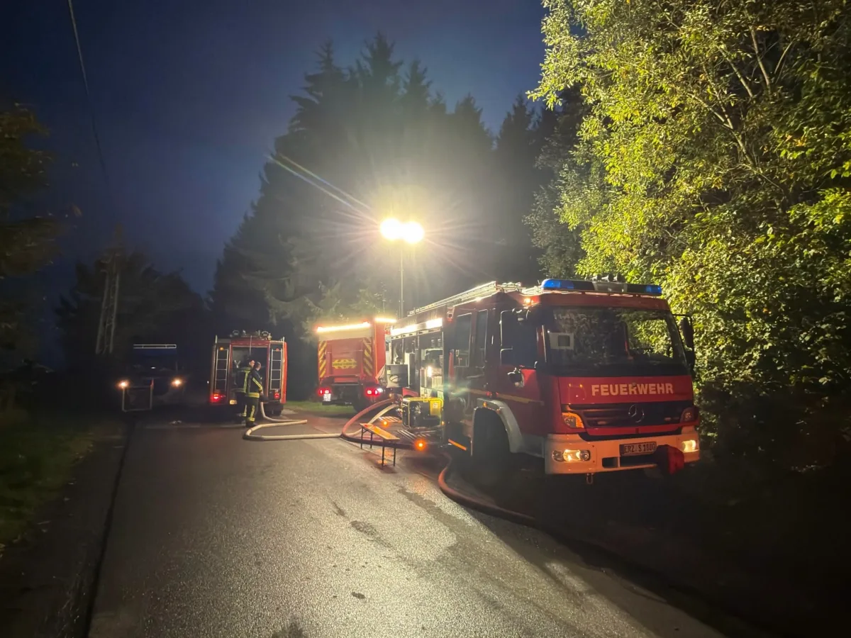 Zwei rote Feuerwehrautos stehen bei Nacht mit eingeschaltetem Licht auf einer Landstraße. Ein Feuerwehrmann ist ebenfalls zu sehen.