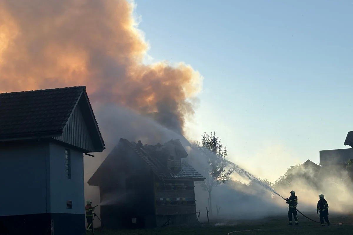 Zwei Feuerwehrleute stehen vor einem stark rauchenden Gebäude und sprühen Wasser aus Schläuchen auf die Flammen. Der Himmel ist leicht bewölkt, die Sonne scheint tief zu stehen.