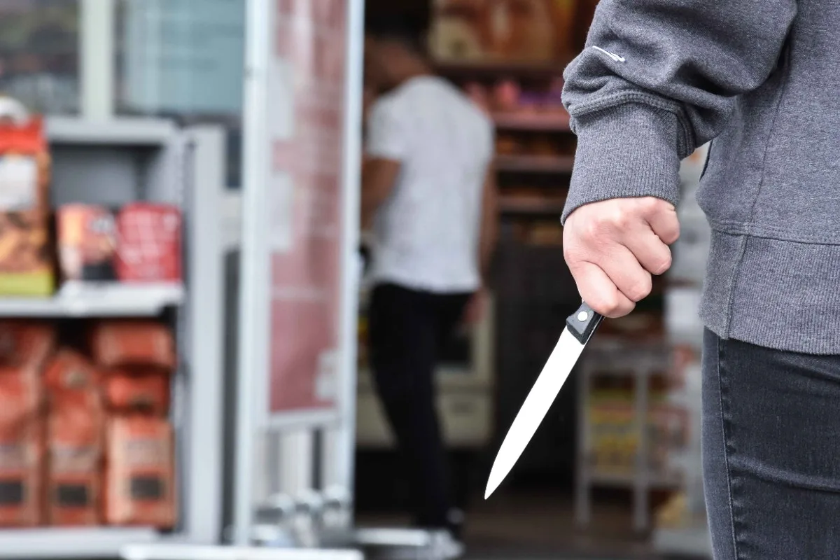 Eine Person in dunkler Kleidung hält ein Messer nahe der Tür eines Geschäfts. Im Hintergrund befindet sich ein weiterer unscharfer Mensch.