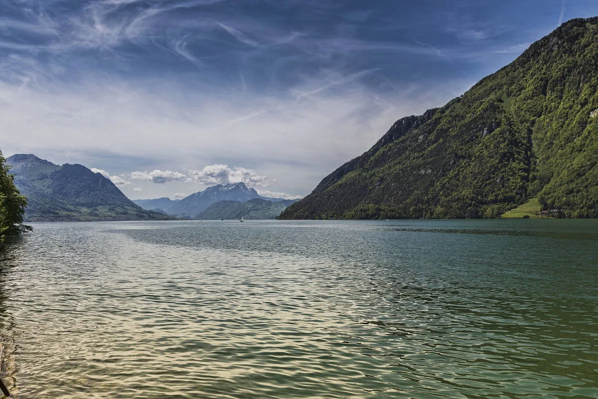 Ein weitläufiger See erstreckt sich vor bewaldeten Bergen unter einem klaren blauen Himmel. Im Hintergrund sind einige Wolken zu sehen.