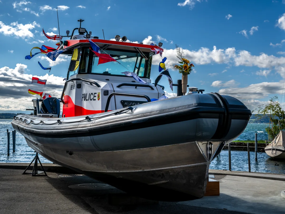 Ein festlich geschmücktes Polizeiboot steht am Ufer. Es ist mit bunten Fahnen dekoriert und der Himmel ist blau mit einigen Wolken.