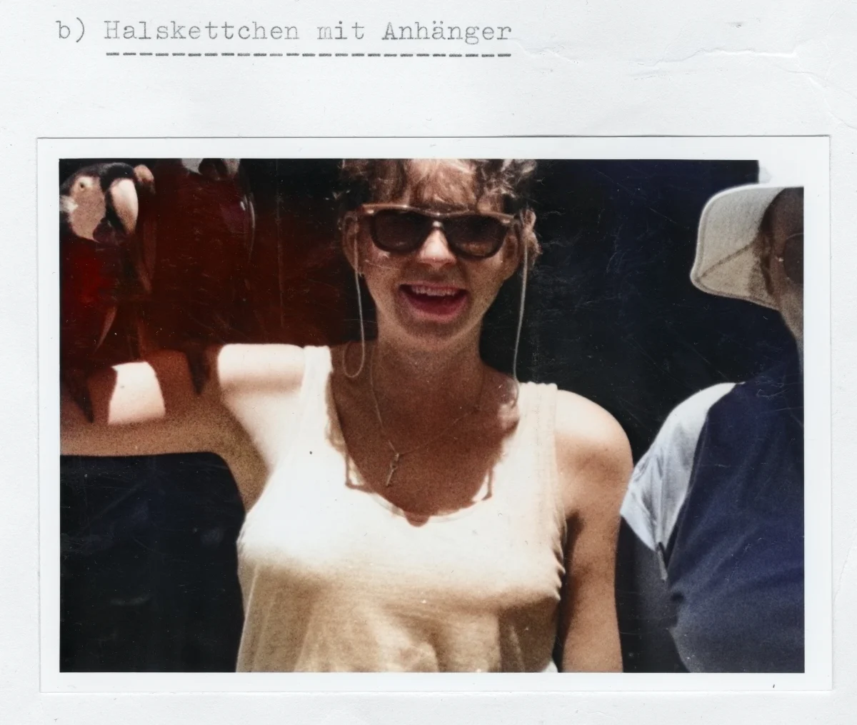 Eine Frau mit Sonnenbrille trägt ein helles Oberteil und eine Kette mit Anhänger. Sie lächelt in die Kamera, neben ihr sind Teile eines Papageis sichtbar.