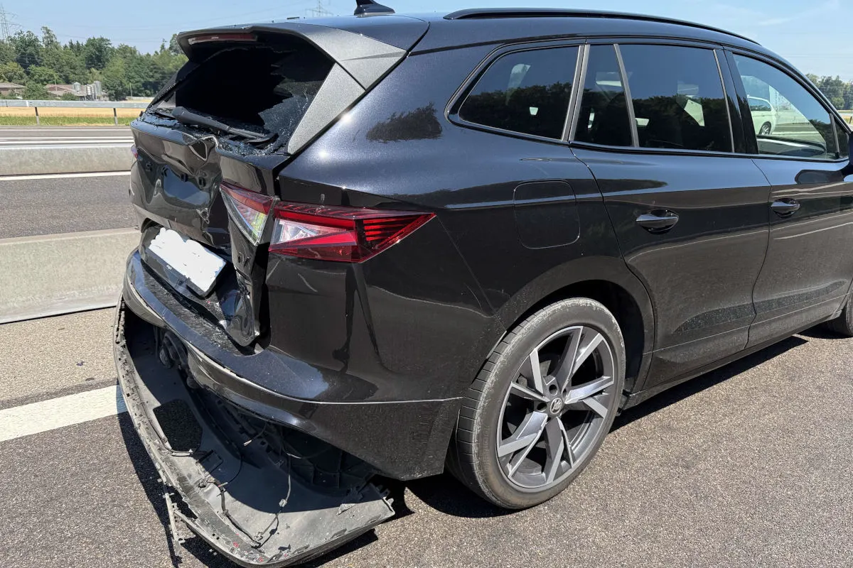 Ein schwarzer Combi steht auf der Autobahn mit stark beschädigtem Heck und abgerissener Stoßstange. Die Autobahn ist an einem sonnigen Tag befahren, umliegende Bäume sind zu sehen.