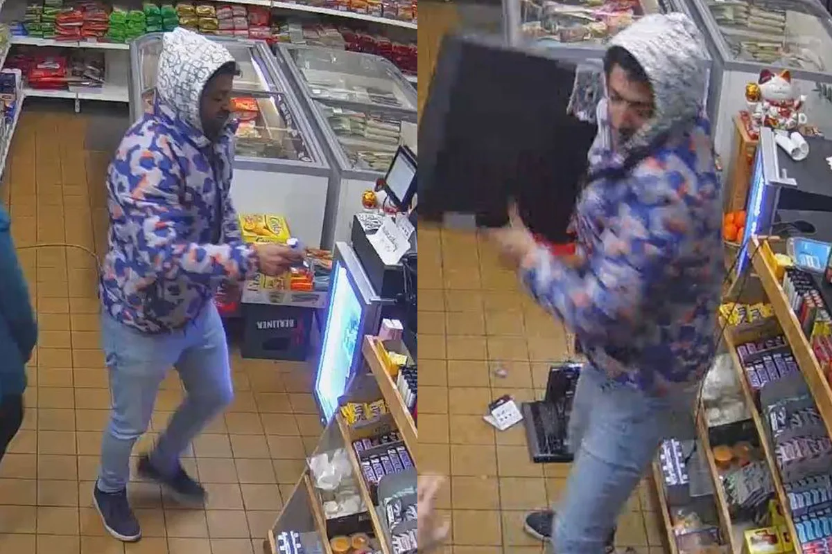 Ein Mann in bunter Jacke steht an der Kasse eines Geschäfts mit einem Gegenstand in der Hand. Im Hintergrund sind Regale mit Waren zu sehen.