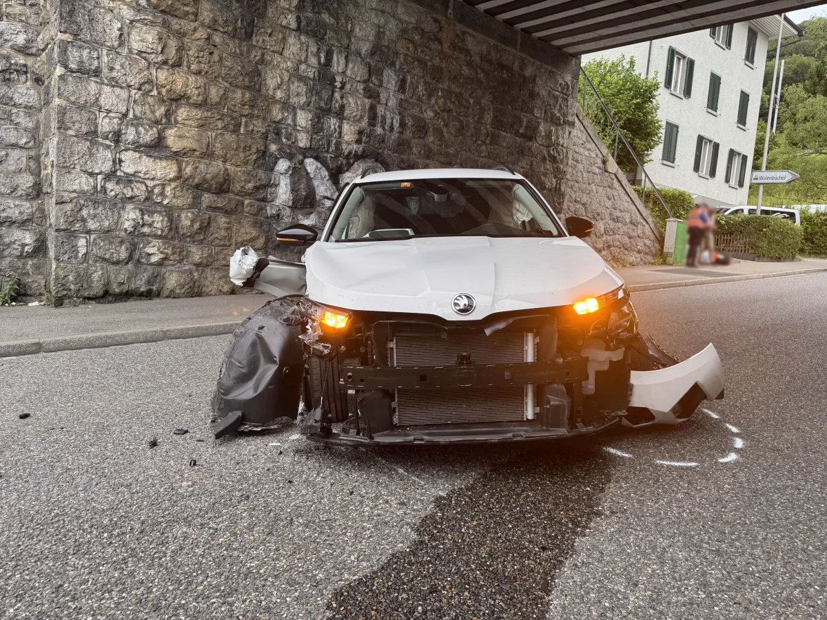 Ein beschädigtes weißes Auto mit eingeschalteten Warnblinkern steht unter einer Brücke vor einer Steinwand. Teile der Stoßstange und Karosserie fehlen oder sind verformt. Im Hintergrund sind zwei Personen unscharf zu erkennen.