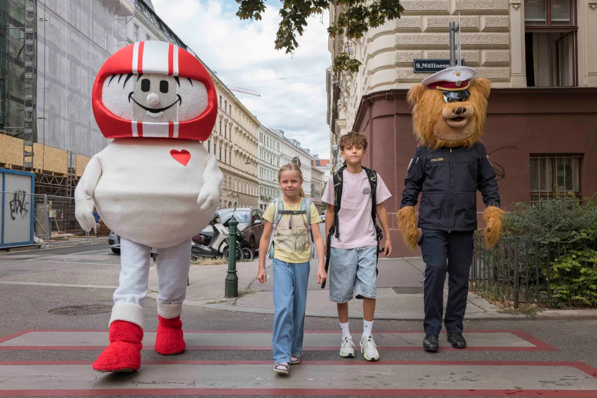 Zwei Kinder gehen auf einem Zebrastreifen, begleitet von einem großen Maskottchen und einem Polizisten in Bärenverkleidung. Die Umgebung ist städtisch.