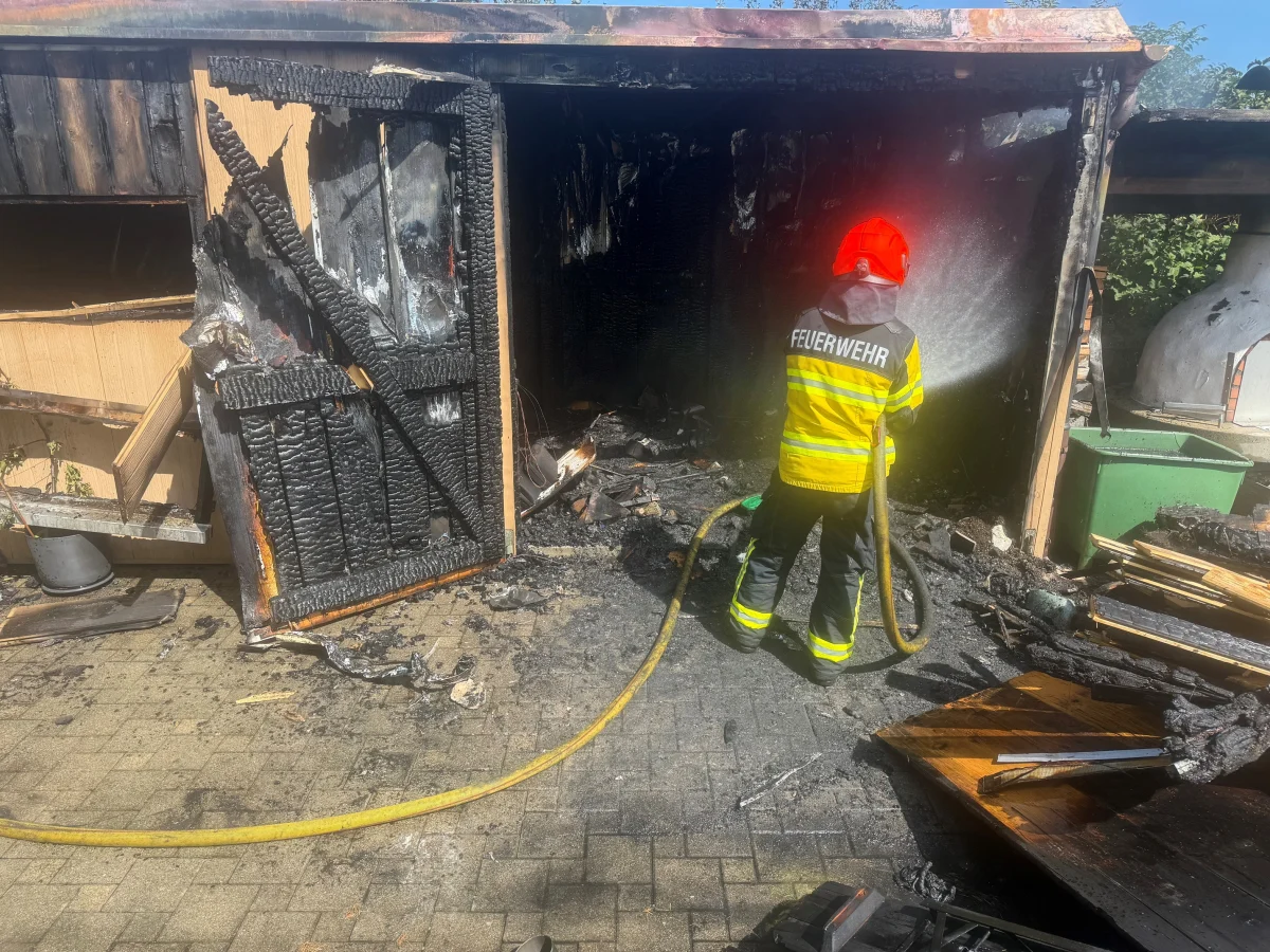 Ein Feuerwehrmann in Einsatzkleidung löscht mit einem Schlauch die verkohlten Reste eines Schuppens. Trümmerteile liegen am Boden.