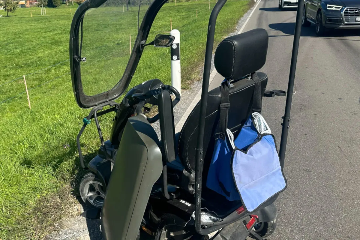 Ein beschädigtes Elektromobil steht am Straßenrand einer Straße, während Fahrzeuge vorbeifahren. Im Hintergrund ist eine Wiese zu sehen.