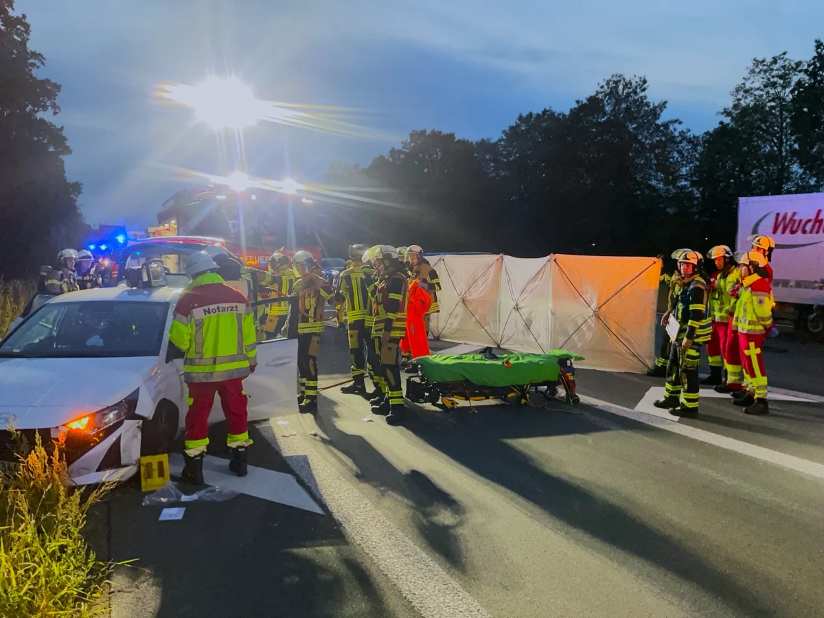 Feuerwehrleute und ein Notarzt stehen bei einem weißen Auto, das an einem Unfall beteiligt ist, während es dämmert. Eine Trage steht bereit.