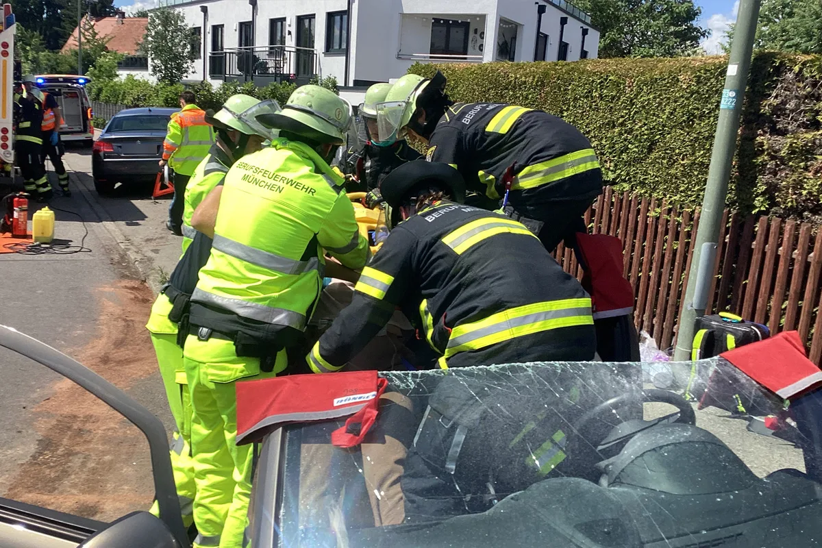 Mehrere Feuerwehrleute der Berufsfeuerwehr München führen eine Rettungsaktion an einem Auto durch, das auf einer Straße steht. Der Fokus liegt auf der Rettungstätigkeit und der Ausstattung der Einsatzkräfte.