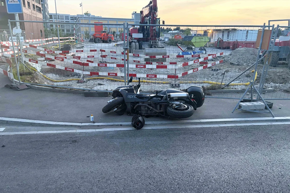Ein schwarzes Motorrad liegt auf der Seite vor einer mit roten und weißen Absperrbändern gesicherten Baustelle. Im Hintergrund sind Baugeräte sichtbar.