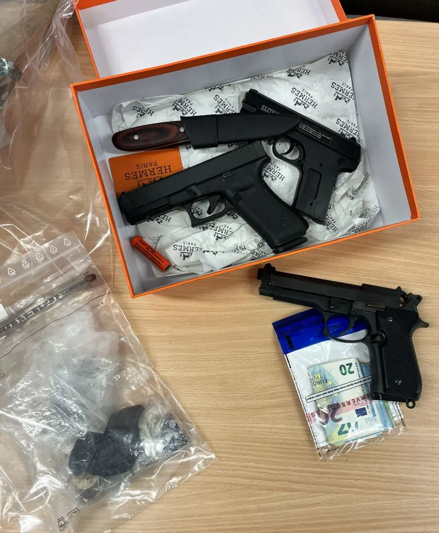 Auf einem Tisch liegen zwei schwarze Pistolen, eine in einer offenen Schachtel und die andere daneben. Daneben sind Bargeld und versiegelte Tüten mit unbekannten Gegenständen sichtbar.