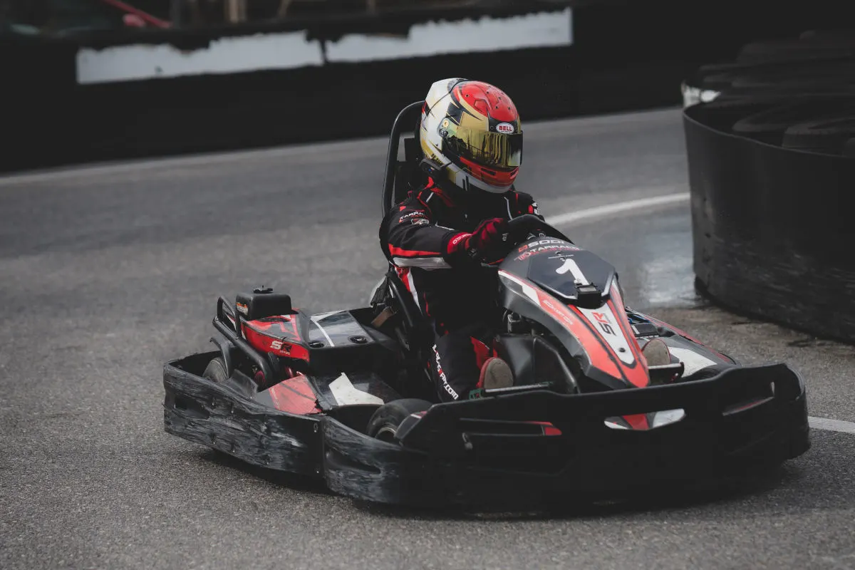 Ein Go-Kart-Fahrer im roten Kartsporthelm fährt auf einer Rennstrecke. Der Fahrer trägt einen schwarzen Rennanzug mit roten Akzenten.