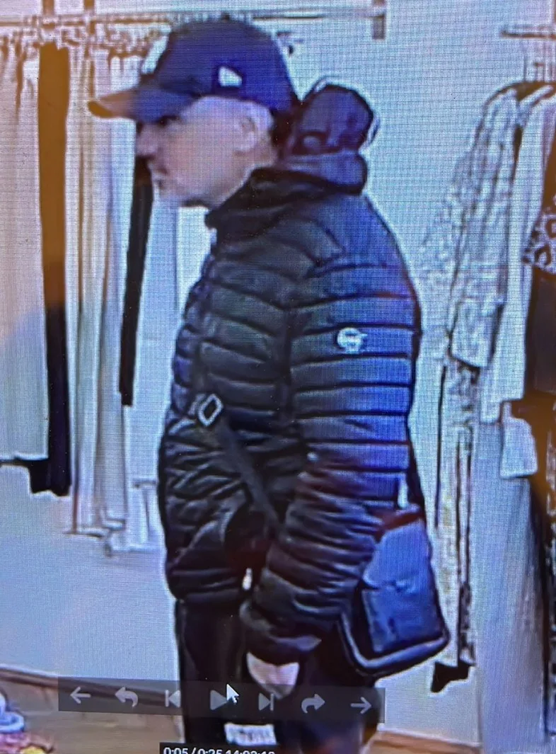 Ein Mann mit schwarzer Jacke und Mütze steht seitlich in einem Geschäft. Im Hintergrund hängen Kleidungsstücke.