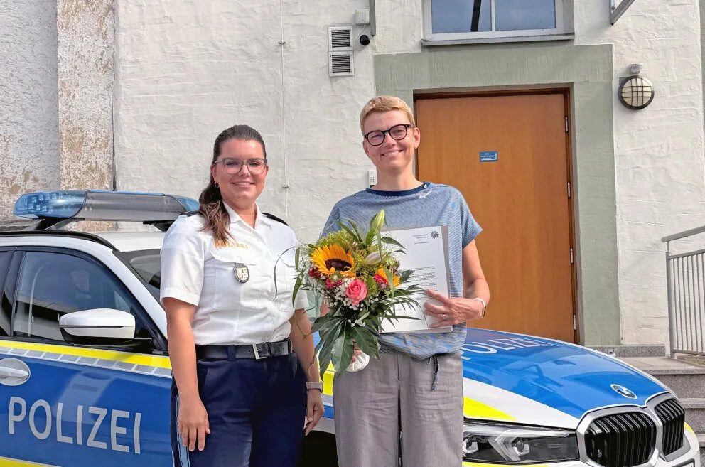 Eine Polizeibeamtin steht neben einer Frau mit Blumen und einer Urkunde vor einem Polizeiwagen.