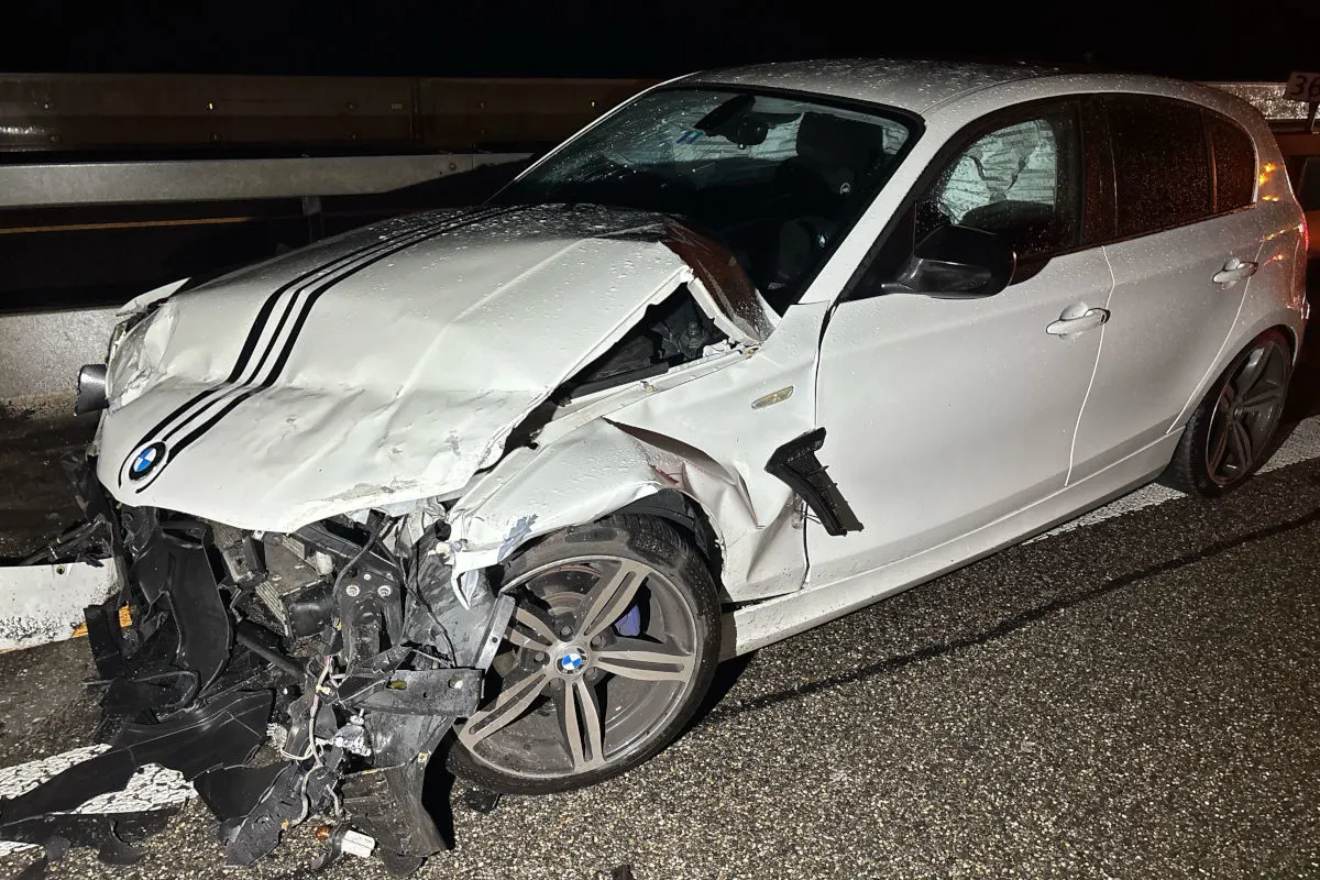 Das Bild zeigt die stark beschädigte Front eines weißen BMWs mit kaputter Stoßstange und Motorhaube. Das Auto steht auf einer Autobahn bei Nacht unter einer Straßenlaterne, und es sind deutliche Schäden an der rechten Vorderseite zu erkennen.