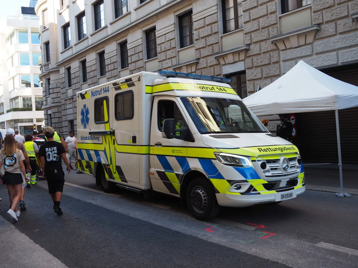 Ein weißer Rettungswagen mit blauen und gelben Streifen parkt in einer engen Stadtstraße neben einem Wohngebäude. Einige Personen gehen die Straße entlang.