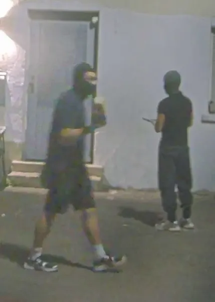Zwei maskierte Personen stehen bei Nacht vor der Tür eines Gebäudes. Eine Person hält einen Gegenstand.